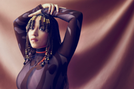 Kaia Kater poses in front of a brown fabric background, looking at the camera with her arms draped over her head.